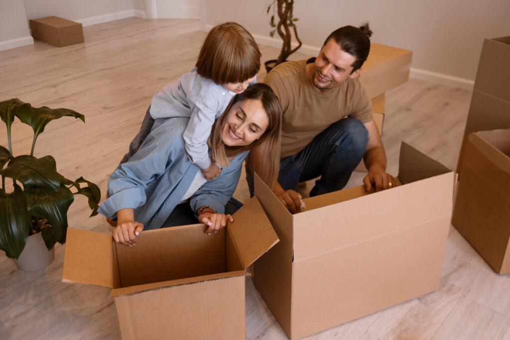 A family of four packing cardboard boxes, with kids playfully tossing stuffed animals into a box.