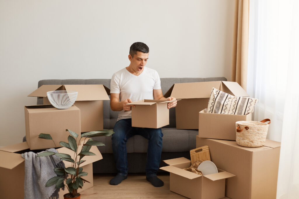 man wearing white casual T-shirt and jeans, sitting on sofa, unpacking boxes before relocating, looking inside box with shocked facial expression.