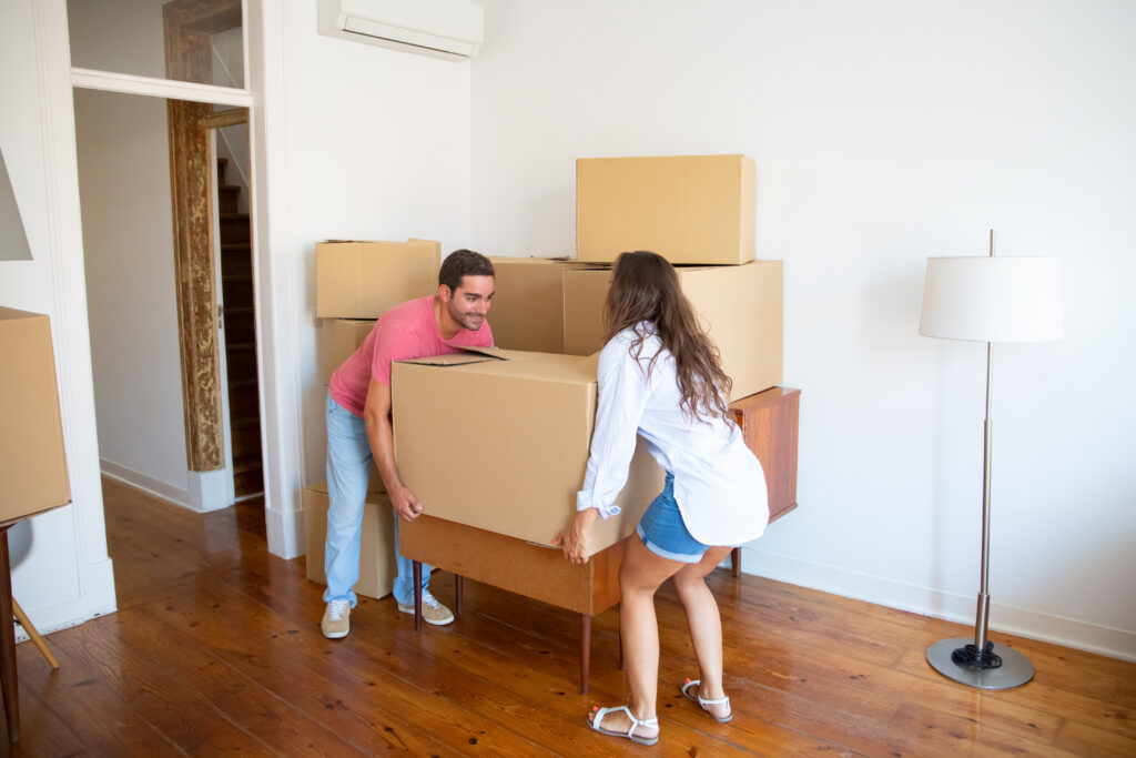 Young family couple moving into new apartment, carrying carton boxes and furniture. Full length. Real estate or property buying concept