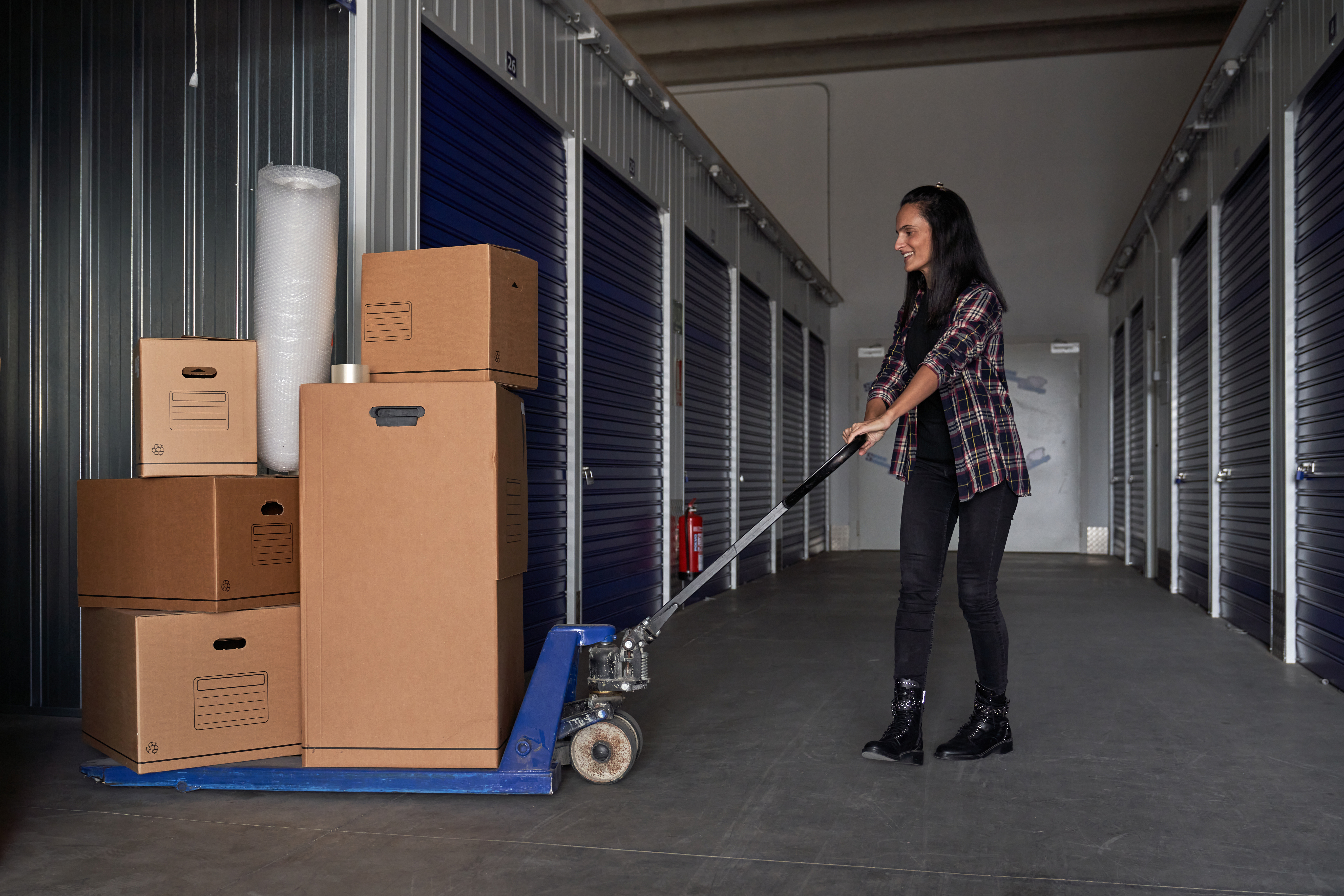 Full body side view of positive female rolling pallet jack with heap of carton boxes near self storage during moving day