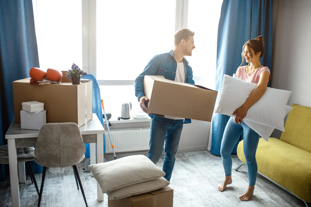 A couple unpacking boxes in a cozy new kitchen with movers in the background.
