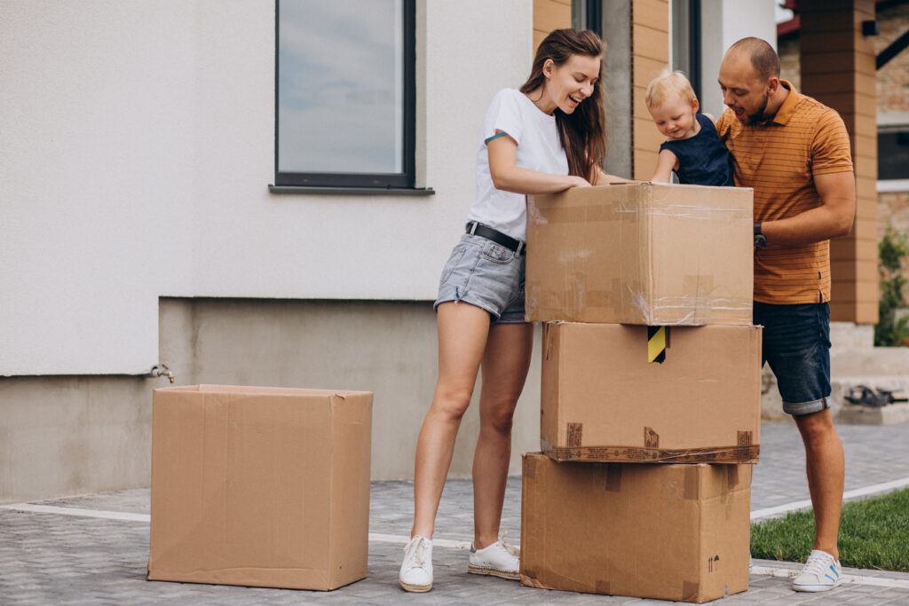 A happy couple standing in front of their new home with moving boxes nearby.
